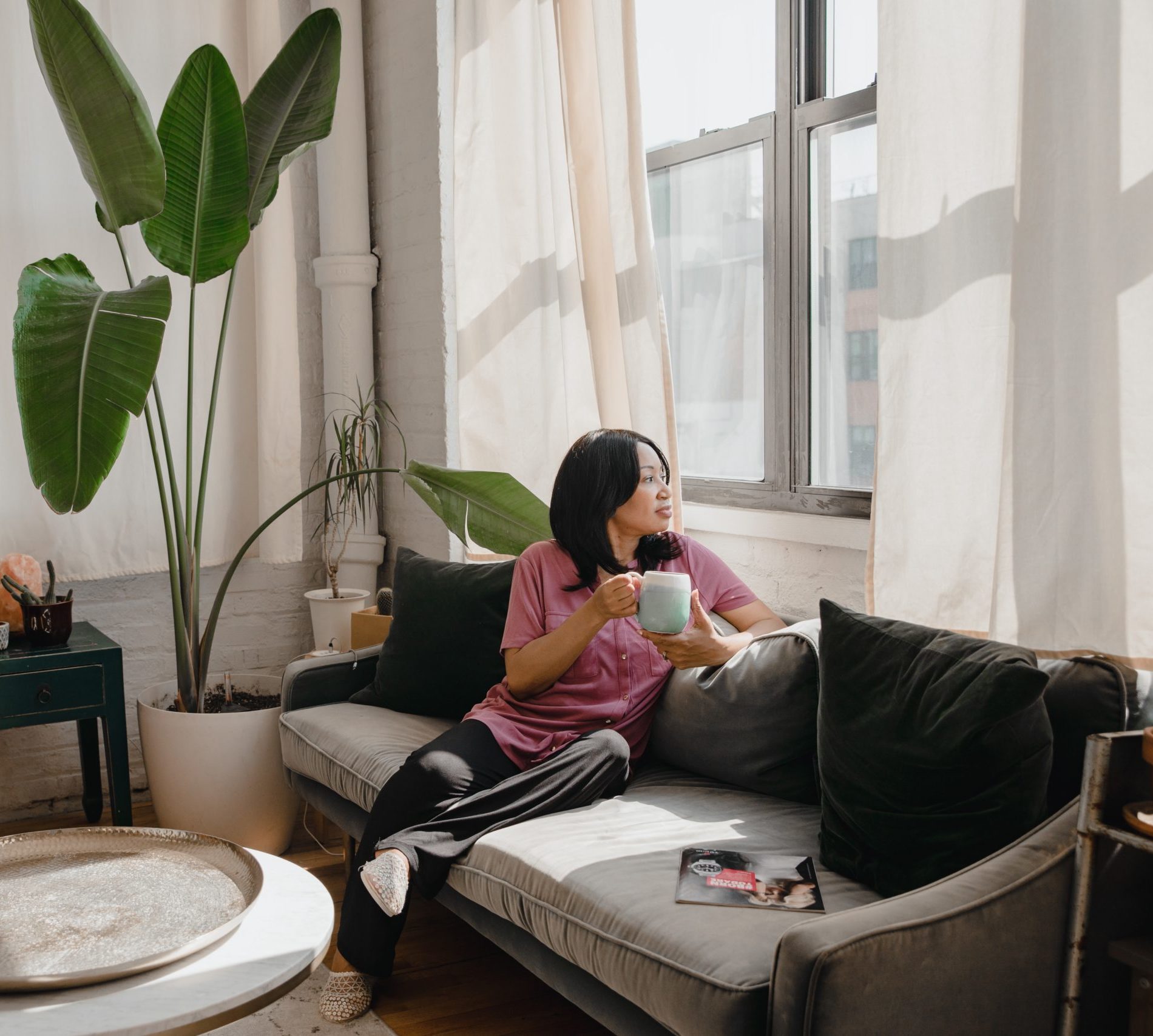 Woman sitting on couch drinking a cup of coffee and staring out of window.